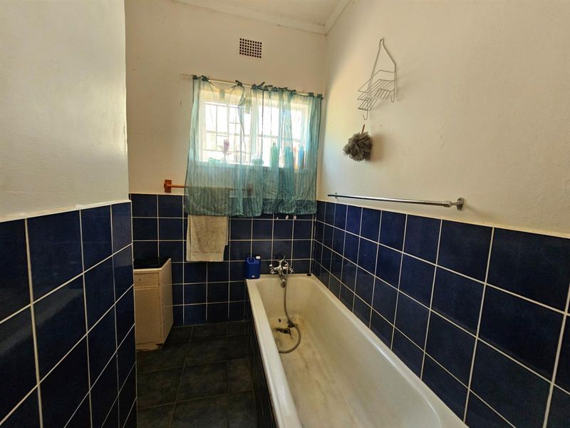 A dated bathroom featuring dark blue wall tiles, a standard bathtub with handheld shower mixer, and a window with security bars.