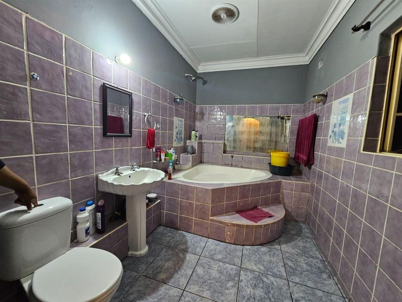 A bathroom featuring a corner bathtub with tiled surround, pedestal basin, and toilet, finished with purple wall tiles and grey floor tiles.