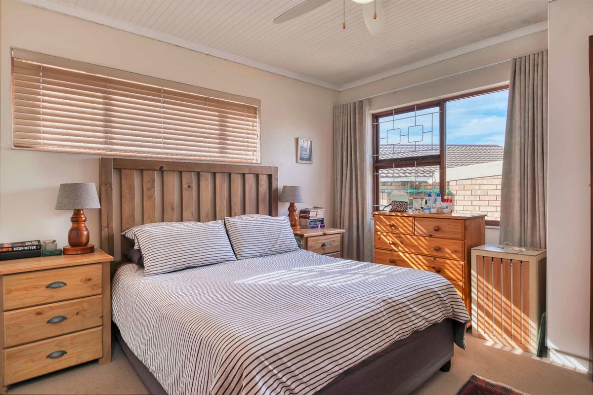A well-lit bedroom featuring a ceiling fan, neutral carpeting, and large windows with blinds, offering a comfortable and functional living space.
