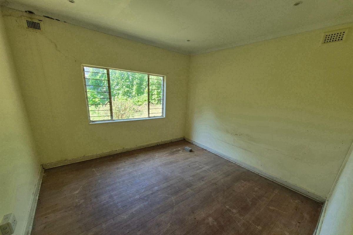 An empty bedroom featuring original timber flooring, large window with security bars, and ceiling ventilation grilles, requiring cosmetic renovation and wall repairs.