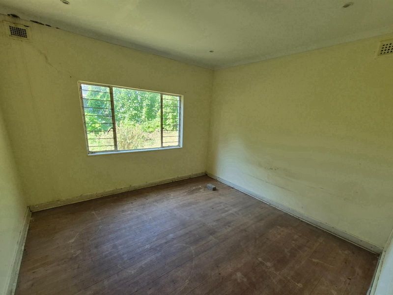 An empty bedroom featuring original timber flooring, large window with security bars, and ceiling ventilation grilles, requiring cosmetic renovation and wall repairs.
