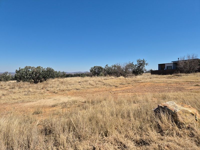 A view of a vacant residential plot featuring natural dry grass, native shrubs, and a large rock formation under a clear blue sky.