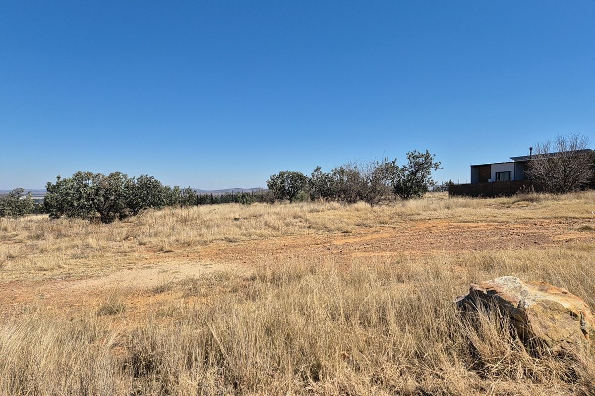 A view of a vacant residential plot featuring natural dry grass, native shrubs, and a large rock formation under a clear blue sky.