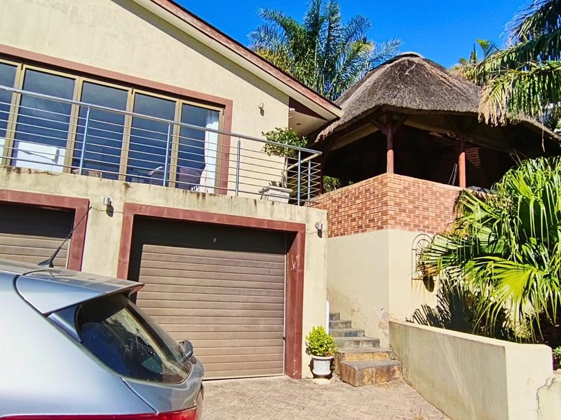Front elevation featuring a double garage, upper balcony with stainless steel balustrade, and a traditional thatched lapa area with paved driveway access.