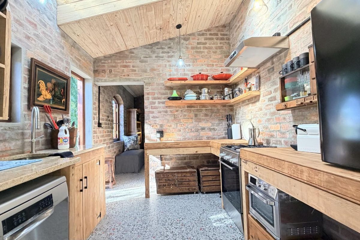 Rustic kitchen featuring exposed brick walls, timber cabinetry, open shelving, a gas stove with extractor fan, and a vaulted timber-clad ceiling.