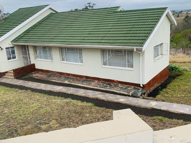 A well-maintained single-story house featuring a green tiled roof, cream-colored exterior walls, and secure metal window grilles, set on a landscaped sloped plot.