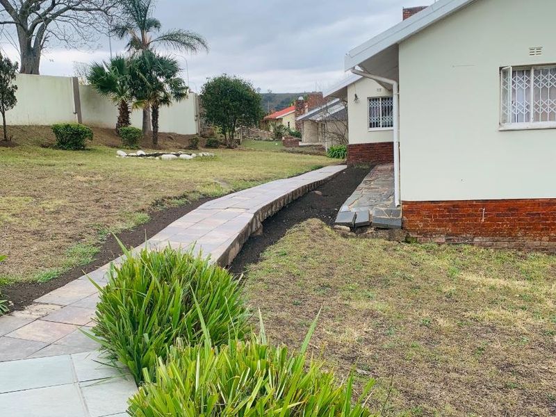 A view of the rear garden featuring a paved walkway, established lawn, and the side elevation of the house with security bars on windows.