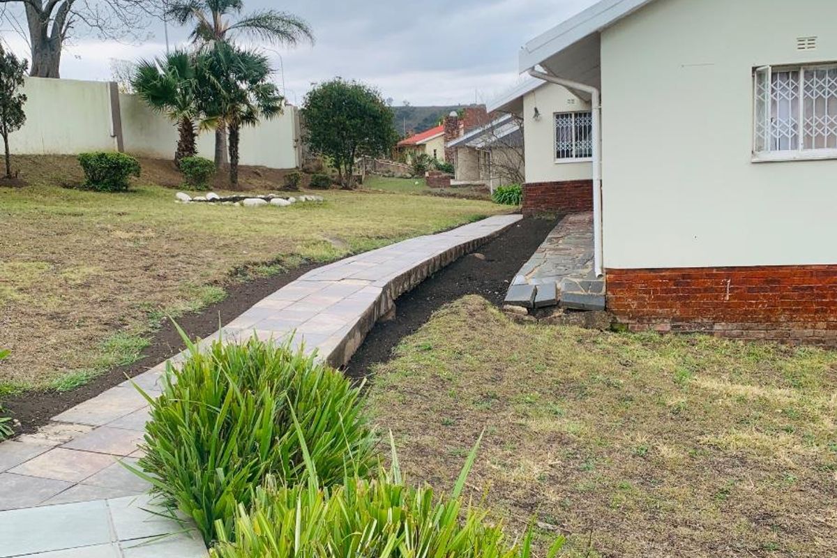 A view of the rear garden featuring a paved walkway, established lawn, and the side elevation of the house with security bars on windows.
