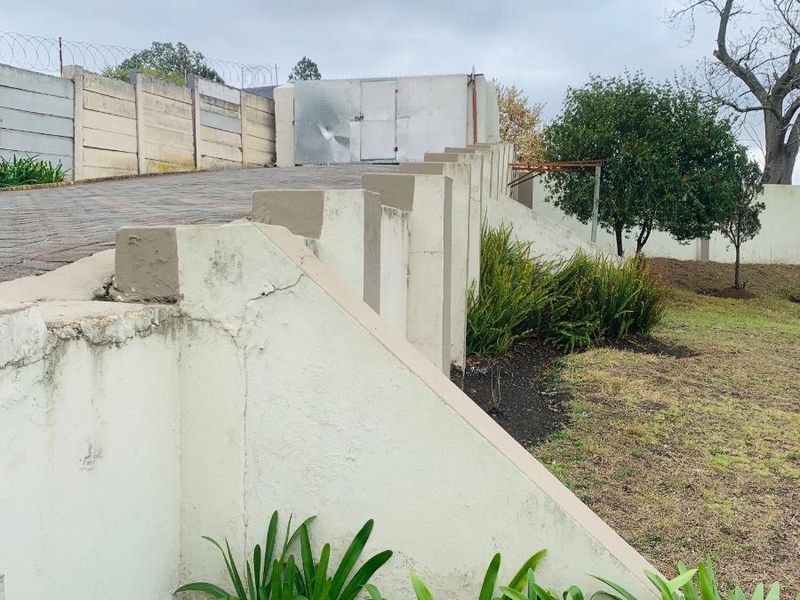 Exterior view of a sloped garden area featuring concrete retaining walls, a paved driveway, and perimeter security fencing.