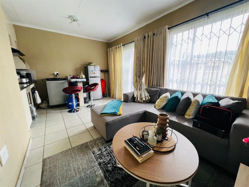 Open-plan living area featuring a compact kitchen with built-in cabinetry and tiled flooring, seamlessly flowing into a comfortable lounge space with large windows.