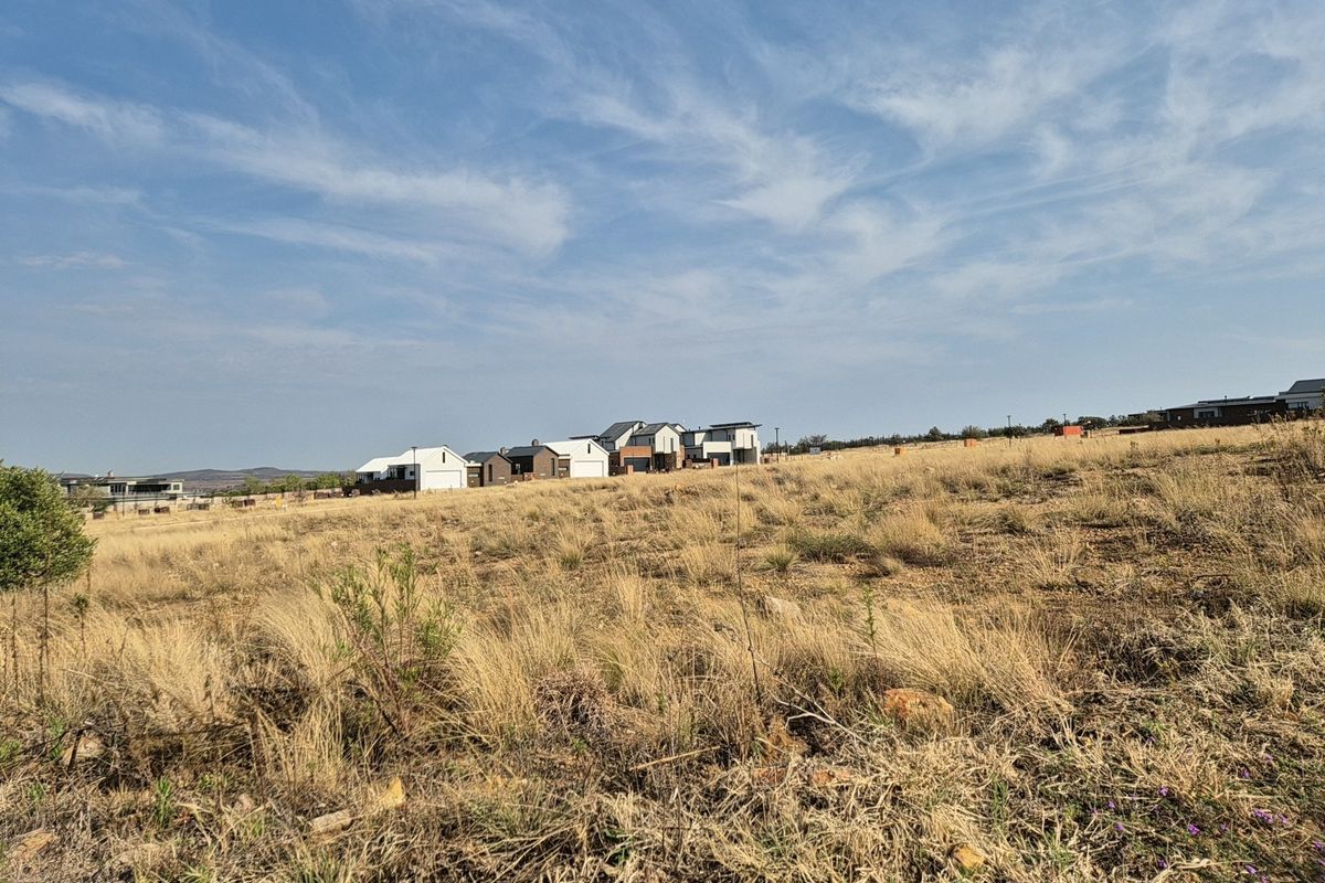 A view of a vacant residential plot featuring natural dry grass vegetation with established residential homes visible in the background.