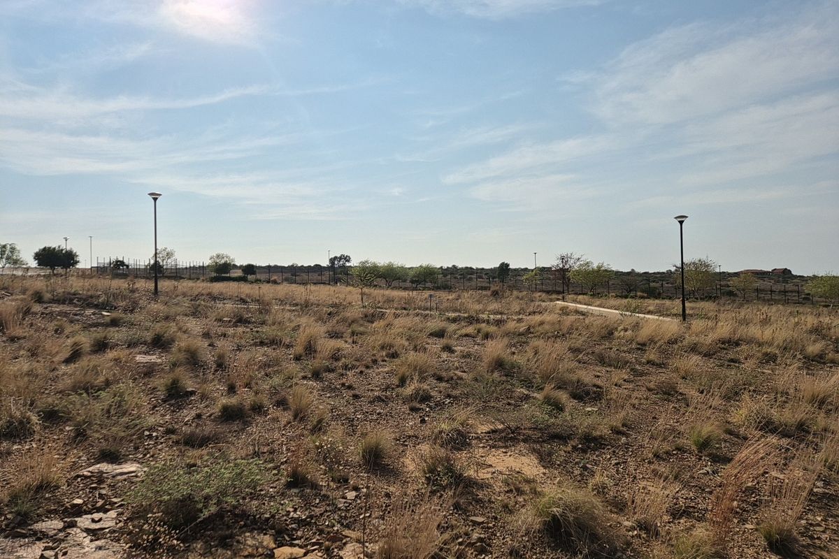 A view of a vacant residential plot featuring natural dry vegetation, perimeter fencing, and installed street lighting poles.