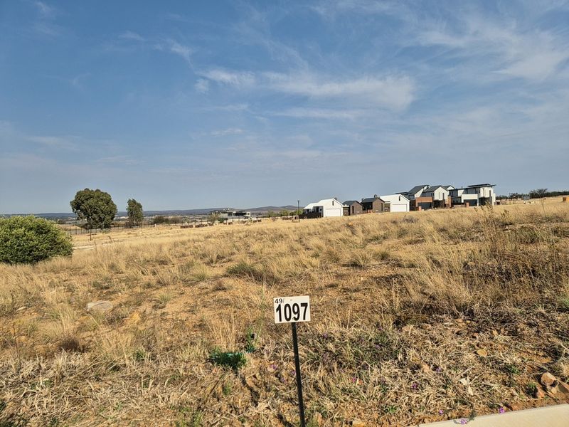 A vacant residential plot featuring natural dry grass vegetation and a clear view of the surrounding estate development.