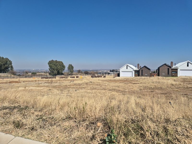 A vacant residential plot of land with dry grass, offering an open outlook towards neighboring modern homes and distant landscape views.