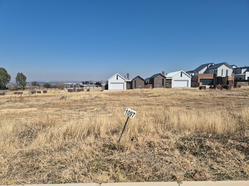 A vacant residential plot of land featuring dry grass and a clear view of neighboring modern homes in a developed estate.