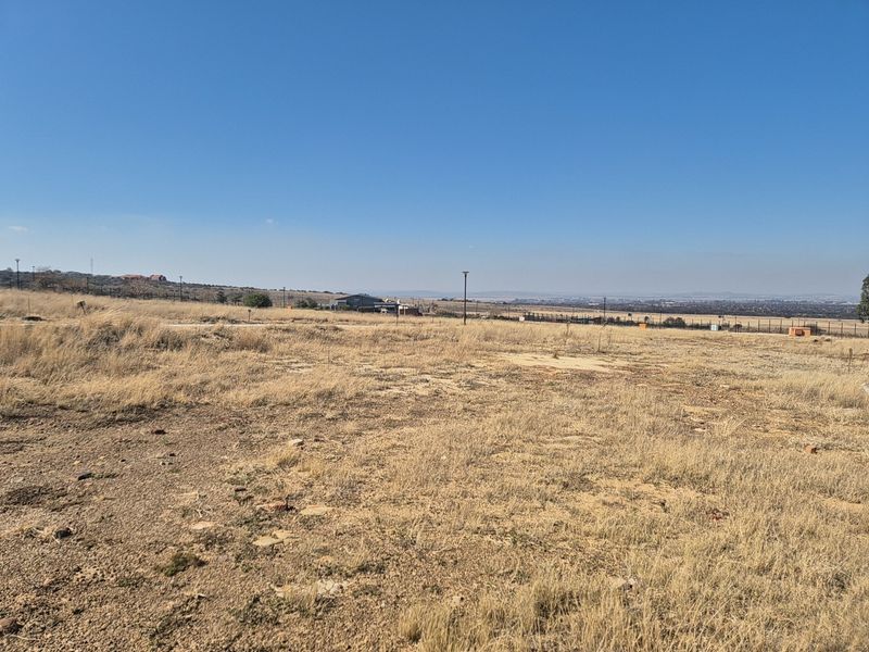 A wide-angle view of a vacant residential plot featuring dry grass, perimeter fencing, and distant landscape views under a clear blue sky.