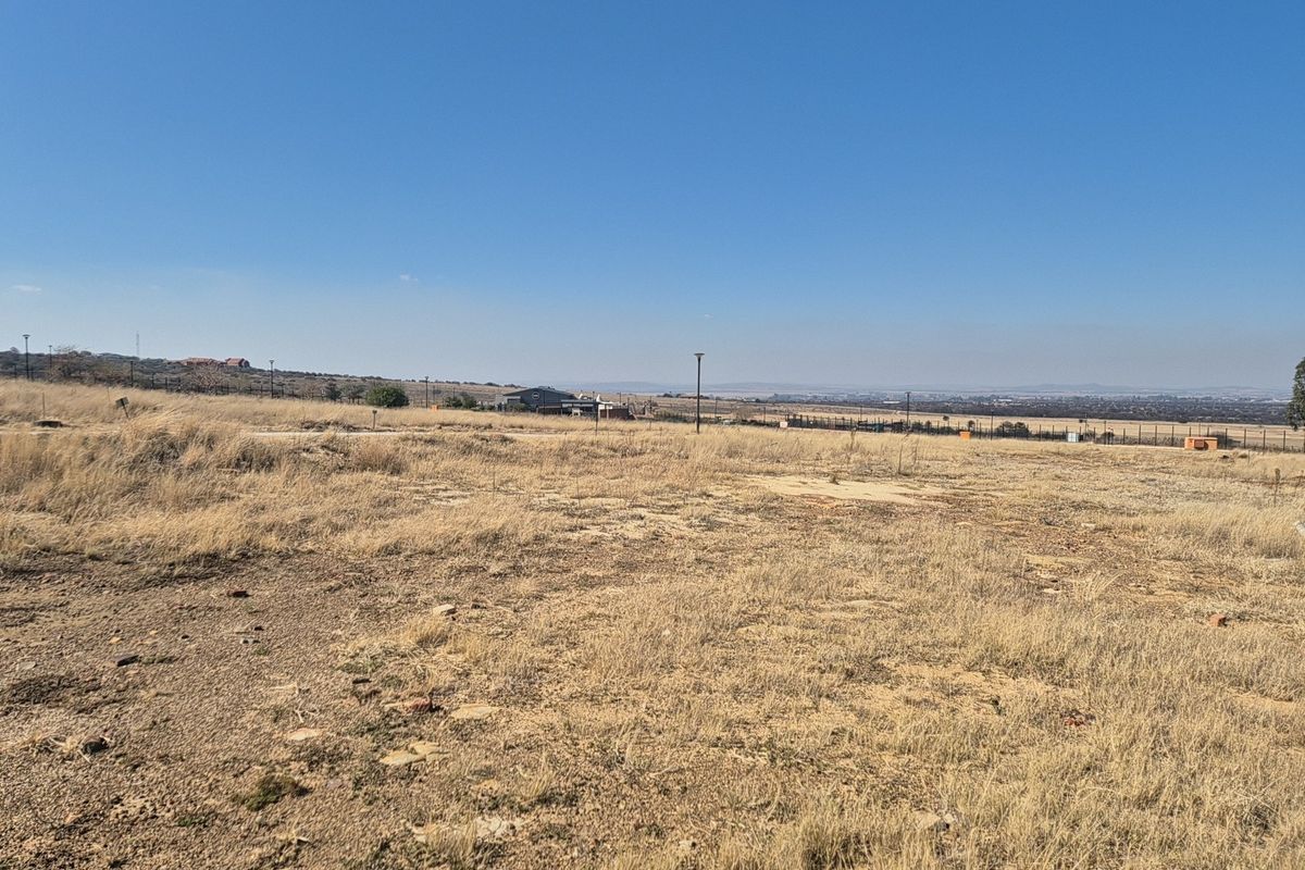 A wide-angle view of a vacant residential plot featuring dry grass, perimeter fencing, and distant landscape views under a clear blue sky.