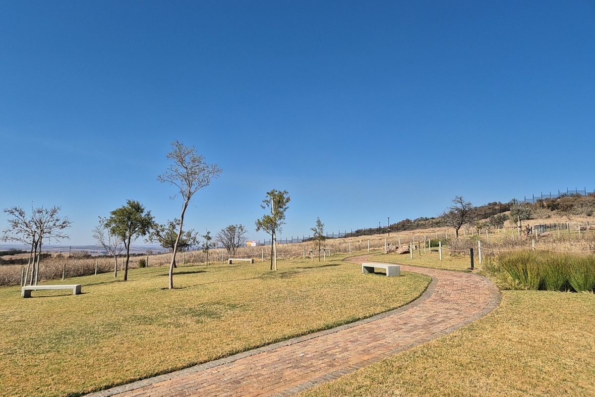 A well-maintained residential garden featuring a brick paved pathway, concrete benches, and established trees set against a clear blue sky.