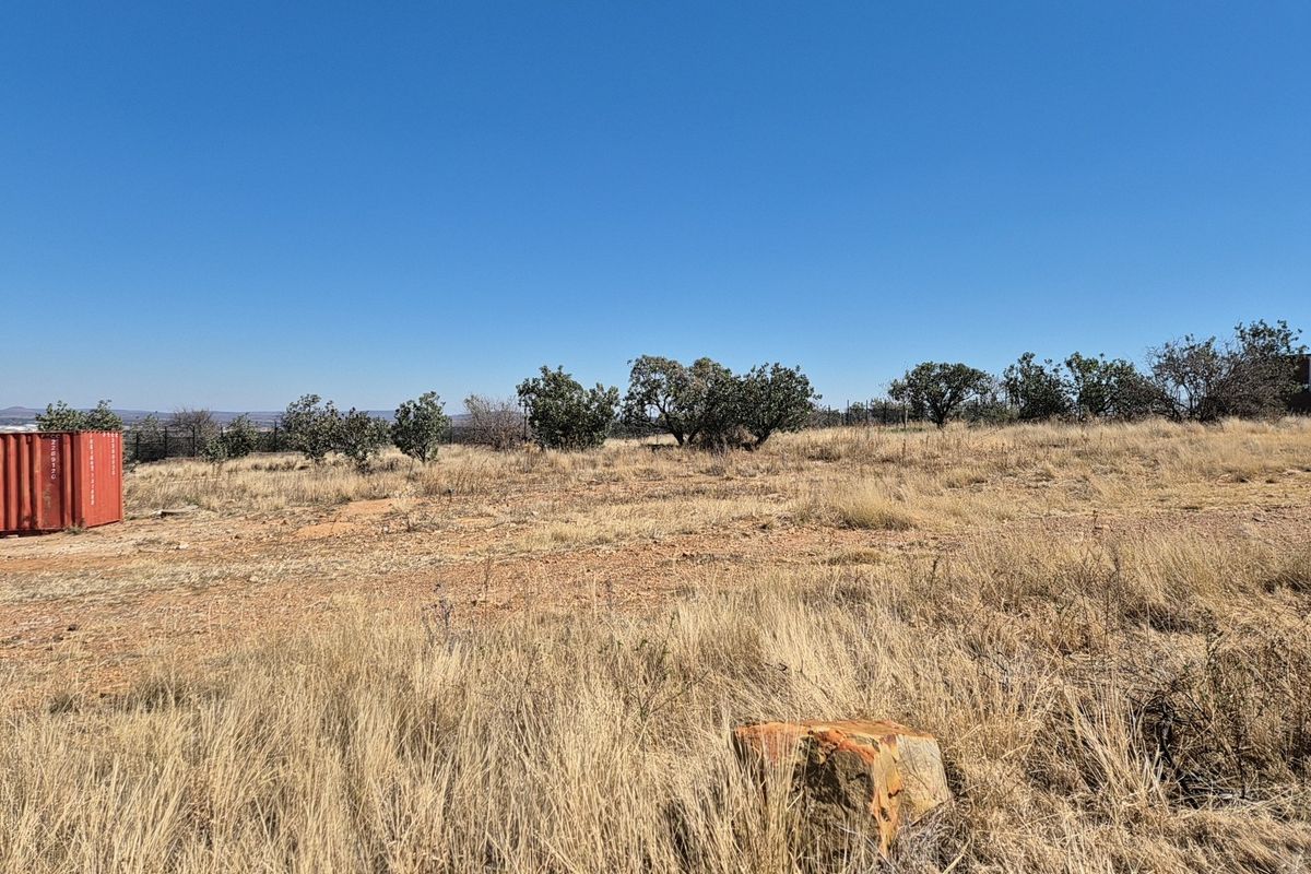 A wide-angle view of a vacant residential plot featuring dry grass, scattered native trees, and perimeter fencing under a clear blue sky.