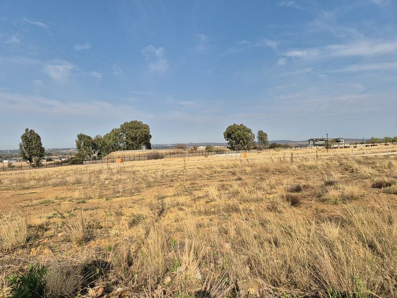 A view of a vacant residential plot featuring dry grass, perimeter fencing, and distant landscape views.
