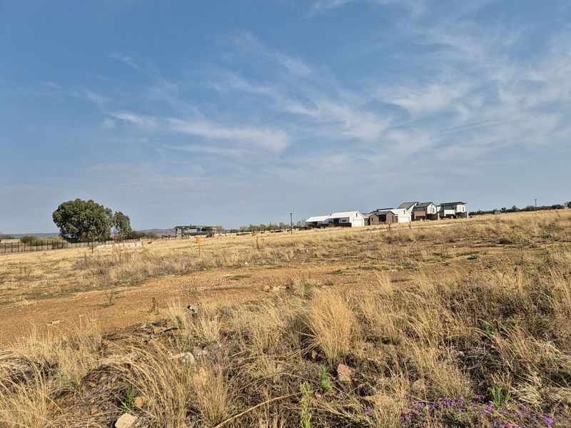 A wide-angle view of a vacant residential plot featuring dry grass and surrounding neighborhood development in the distance.