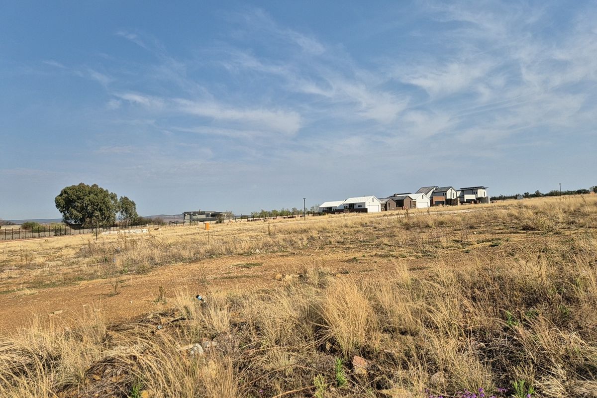 A wide-angle view of a vacant residential plot featuring dry grass and surrounding neighborhood development in the distance.