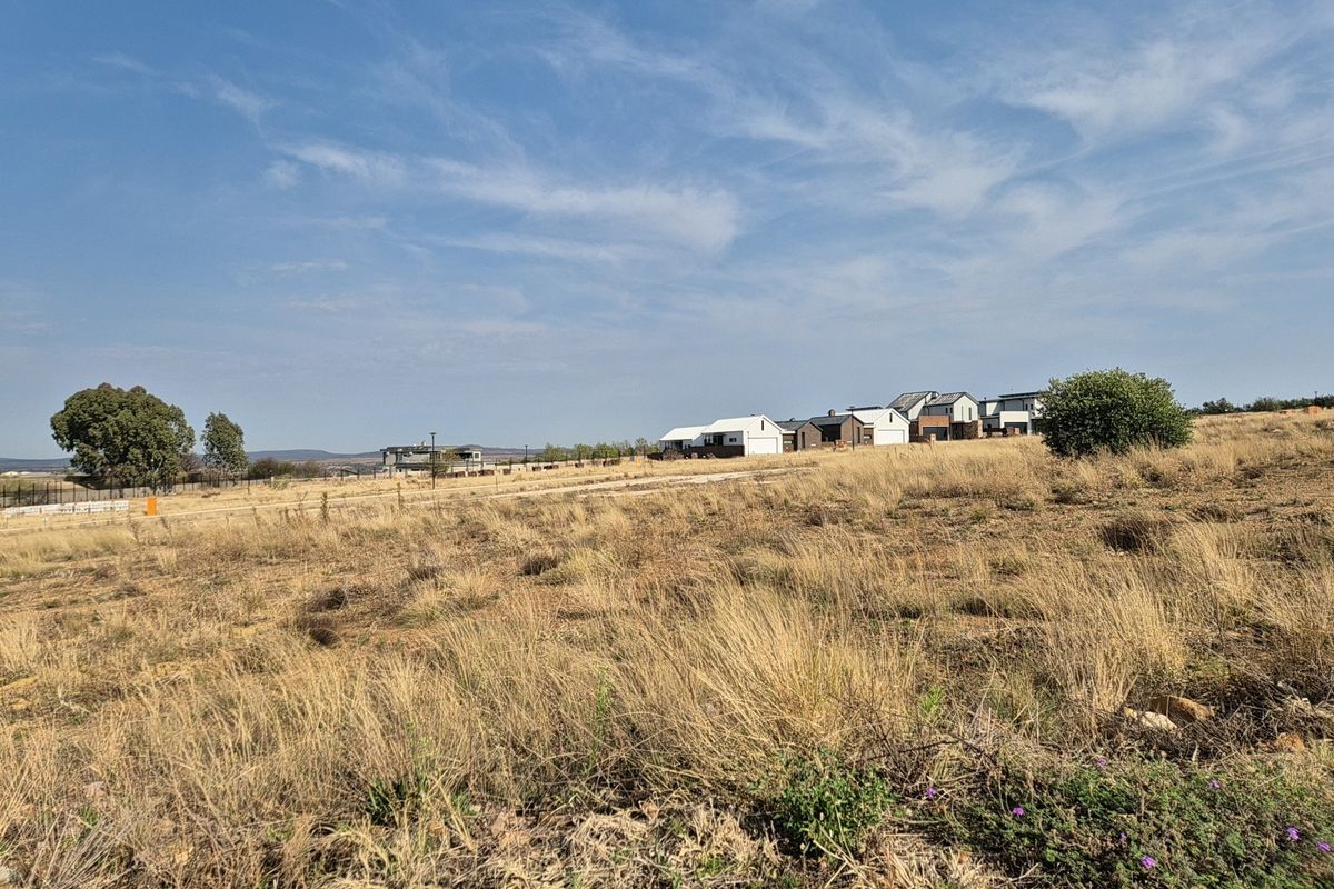 A view of a vacant residential plot featuring dry grass and surrounding neighborhood development in the distance.