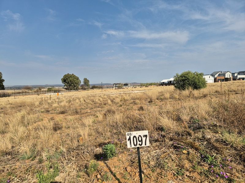 A vacant residential plot featuring natural dry grass landscape, perimeter fencing, and a clear view of the surrounding estate.