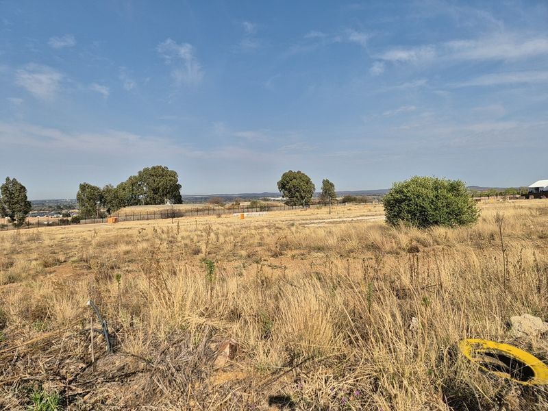 A wide-angle view of a vacant residential plot featuring dry grass, perimeter fencing, and distant landscape views.