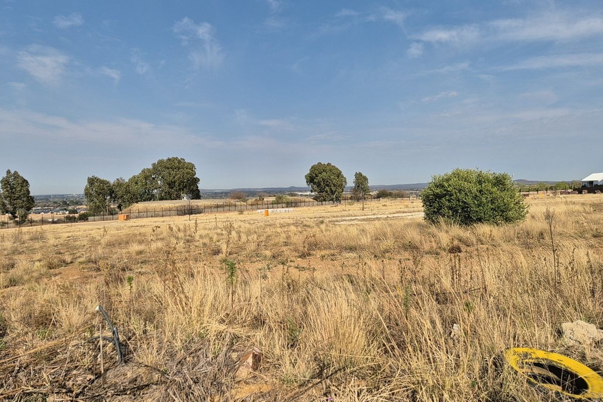 A wide-angle view of a vacant residential plot featuring dry grass, perimeter fencing, and distant landscape views.