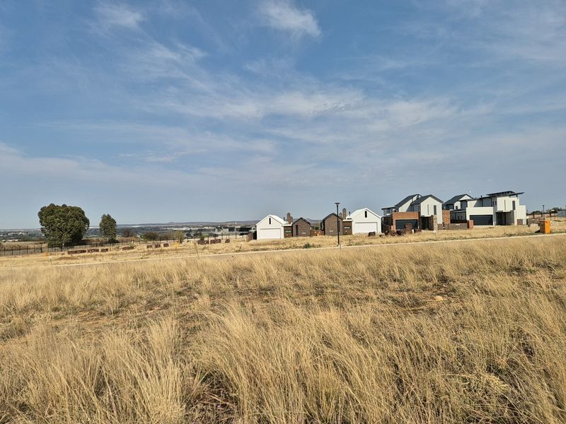 A wide-angle view of an undeveloped residential plot featuring dry grass, overlooking a modern housing estate in the distance.