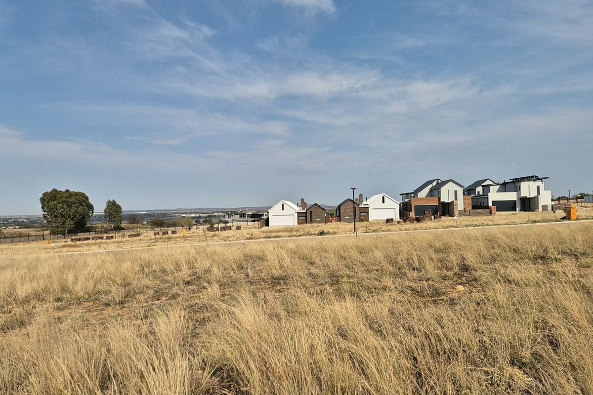 A wide-angle view of an undeveloped residential plot featuring dry grass, overlooking a modern housing estate in the distance.
