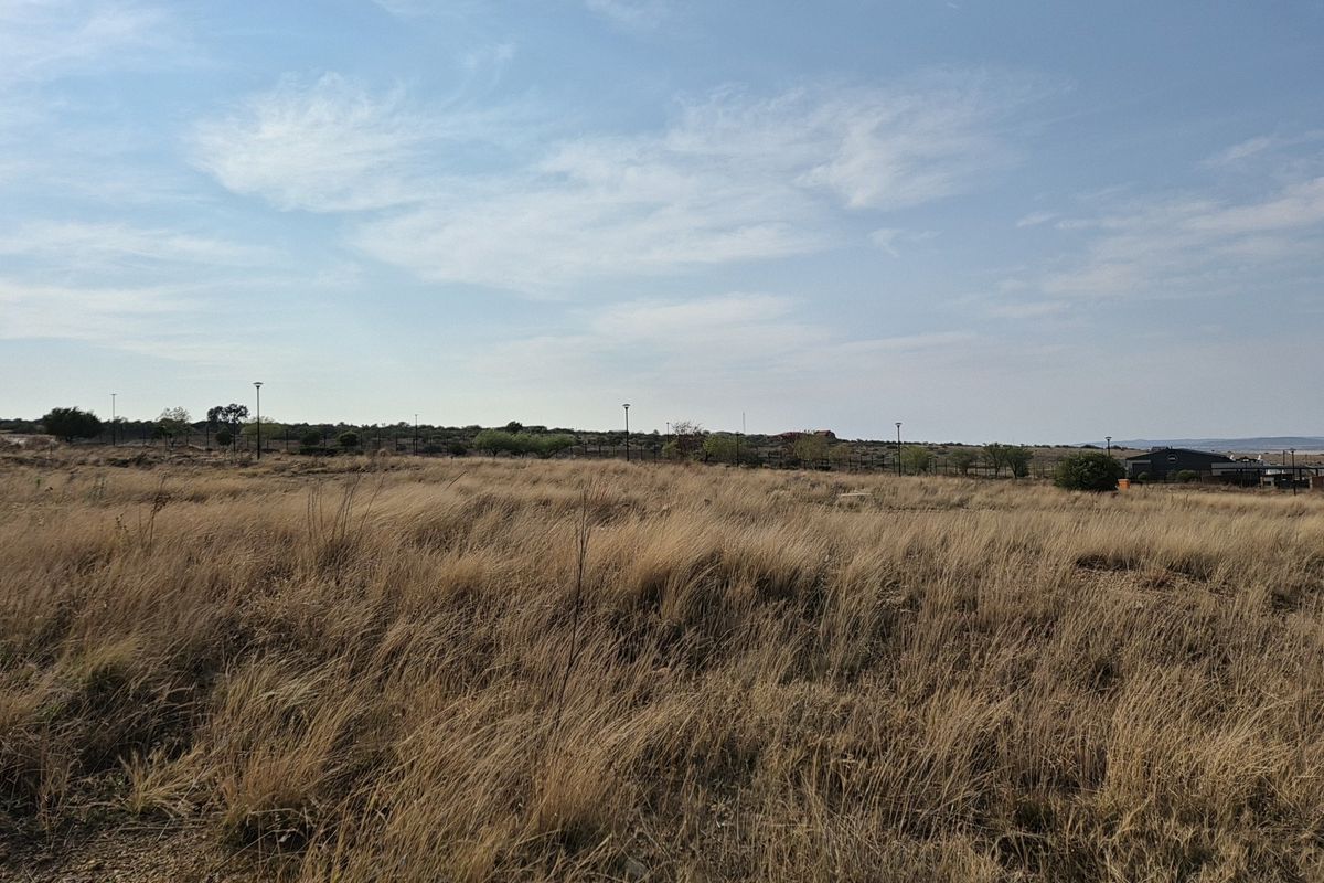 A wide-angle view of a vacant residential plot featuring dry grass, perimeter fencing, and distant street lighting.