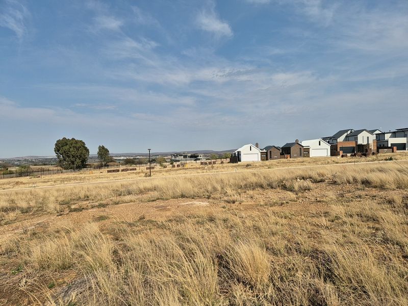 A vacant residential plot featuring dry grass landscape with surrounding neighborhood houses and perimeter fencing visible in the distance.