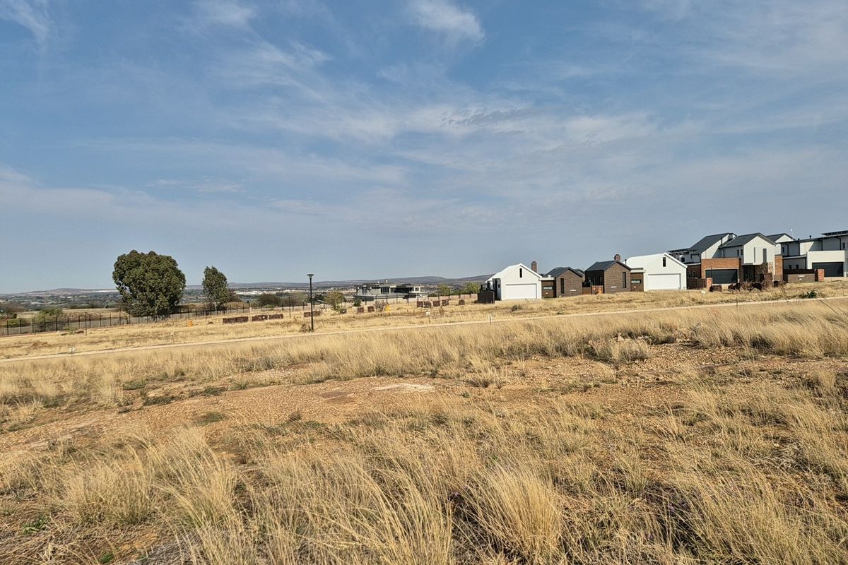 A vacant residential plot featuring dry grass landscape with surrounding neighborhood houses and perimeter fencing visible in the distance.
