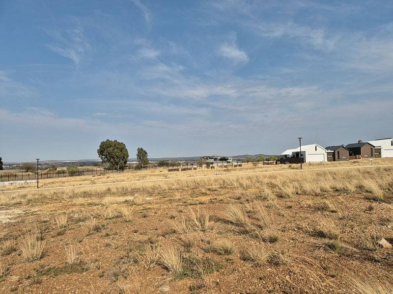 A vacant residential plot featuring dry grass, perimeter fencing, and street lighting, situated in a developing neighborhood with surrounding residential properties.