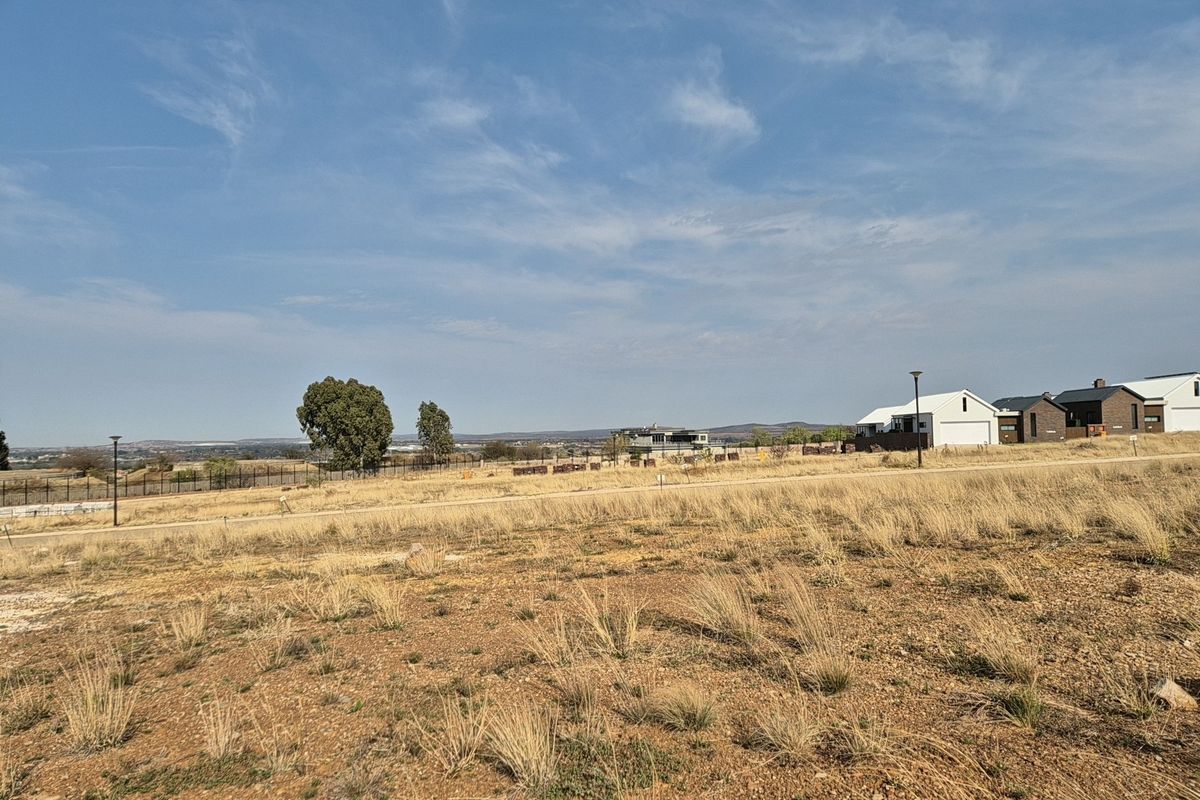 A vacant residential plot featuring dry grass, perimeter fencing, and street lighting, situated in a developing neighborhood with surrounding residential properties.