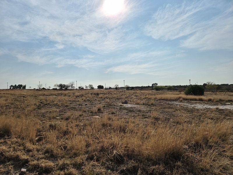 A wide-angle view of a vacant residential plot featuring dry grass, perimeter fencing, and existing street lighting infrastructure.