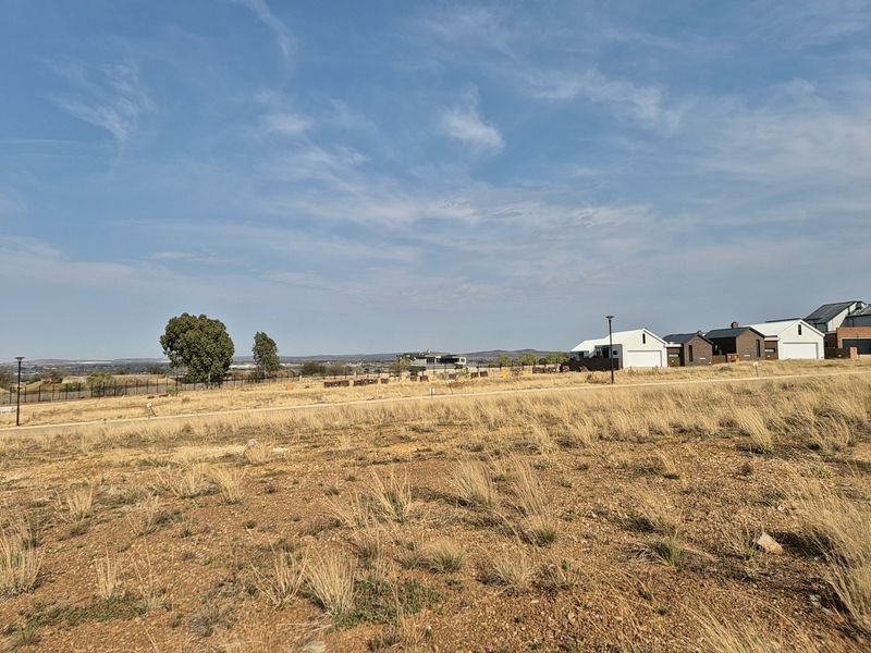 A wide-angle view of a vacant residential plot featuring dry grass, perimeter fencing, and distant neighborhood houses under a clear blue sky.