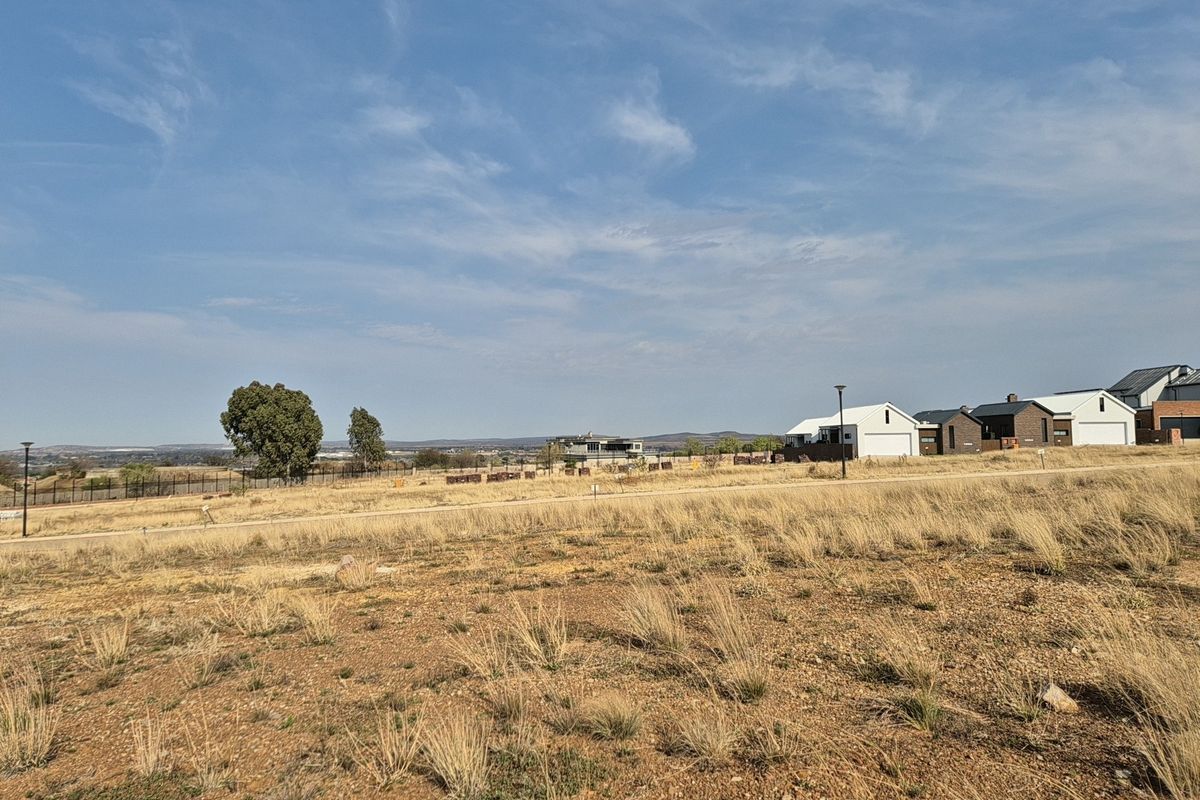 A wide-angle view of a vacant residential plot featuring dry grass, perimeter fencing, and distant neighborhood houses under a clear blue sky.