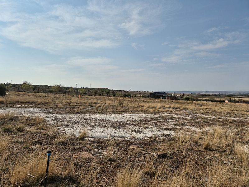 A vacant residential plot featuring natural dry grass terrain, perimeter fencing, and distant views of the surrounding estate landscape.
