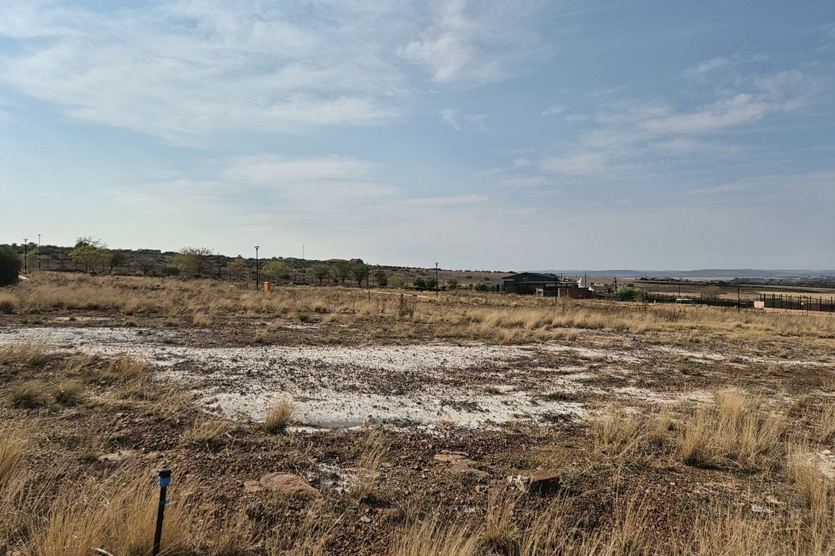 A vacant residential plot featuring natural dry grass terrain, perimeter fencing, and distant views of the surrounding estate landscape.