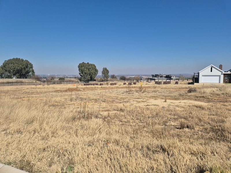 A vacant residential plot of land featuring dry grass, perimeter fencing, and distant views of the surrounding neighborhood.