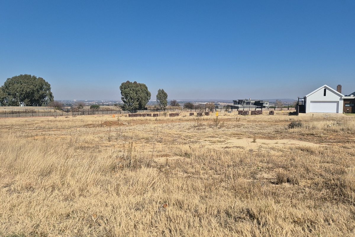 A vacant residential plot of land featuring dry grass, perimeter fencing, and distant views of the surrounding neighborhood.