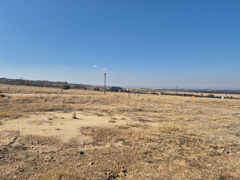 A wide-angle view of a vacant residential plot featuring natural dry grass terrain, perimeter fencing, and street lighting infrastructure.