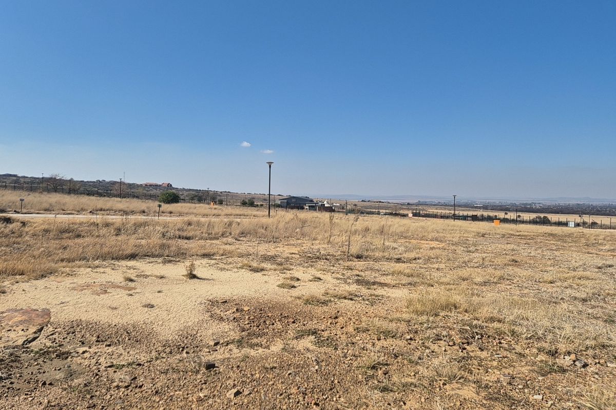 A wide-angle view of a vacant residential plot featuring natural dry grass terrain, perimeter fencing, and street lighting infrastructure.