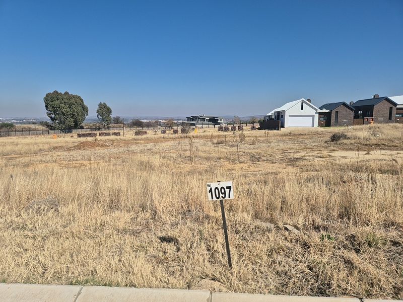 A vacant residential plot of land featuring dry grass, a property marker, and surrounding neighborhood development in the background.
