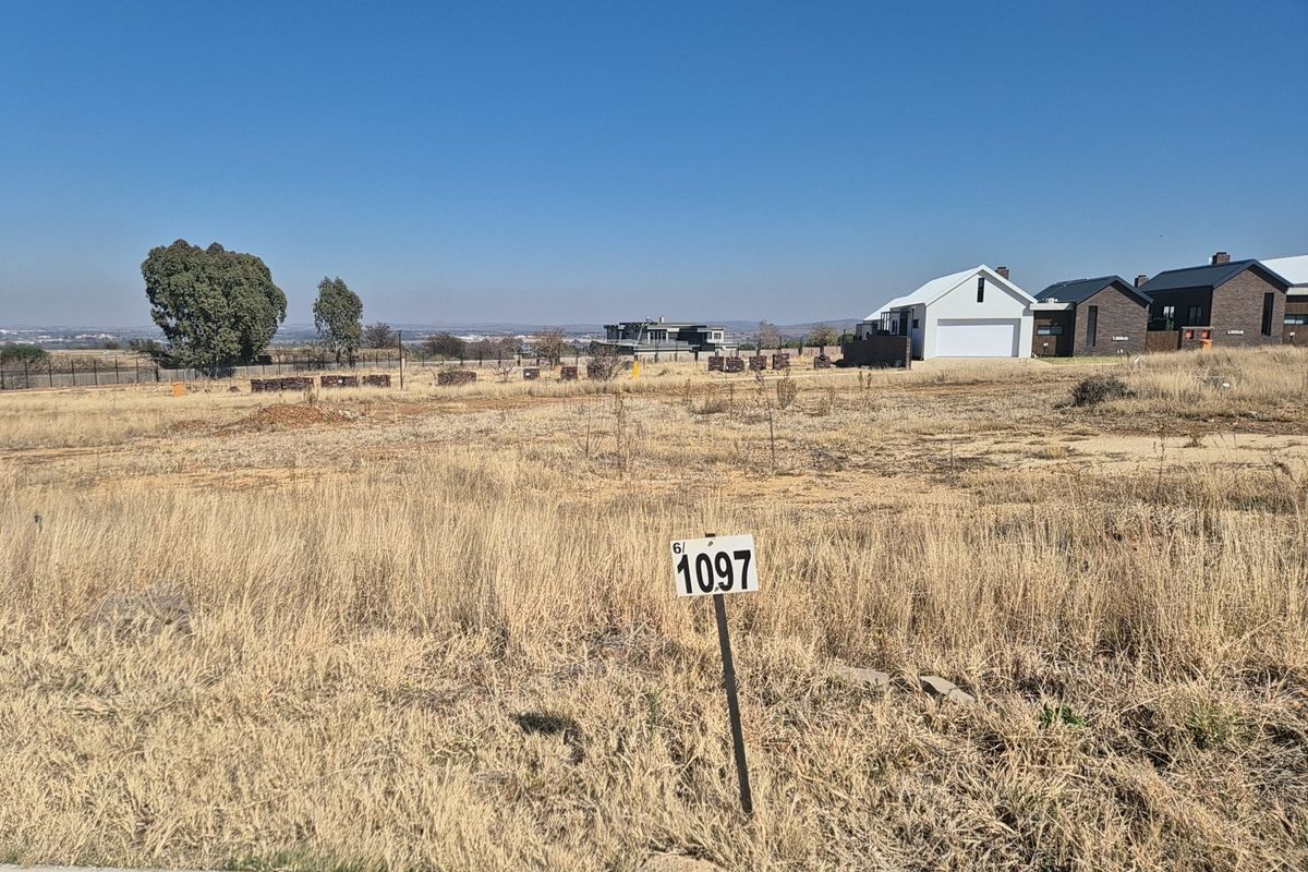 A vacant residential plot of land featuring dry grass, a property marker, and surrounding neighborhood development in the background.