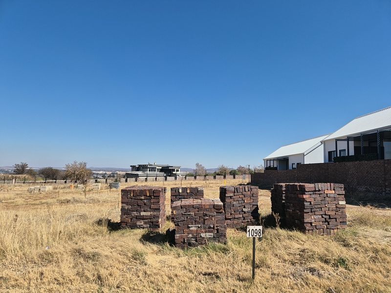 A vacant residential plot featuring dry grass, a boundary wall, and a plot marker, situated in a developed residential estate.
