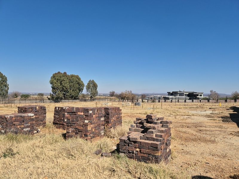 A vacant residential plot featuring dry grass, stacks of construction bricks, and perimeter fencing with distant neighborhood views.